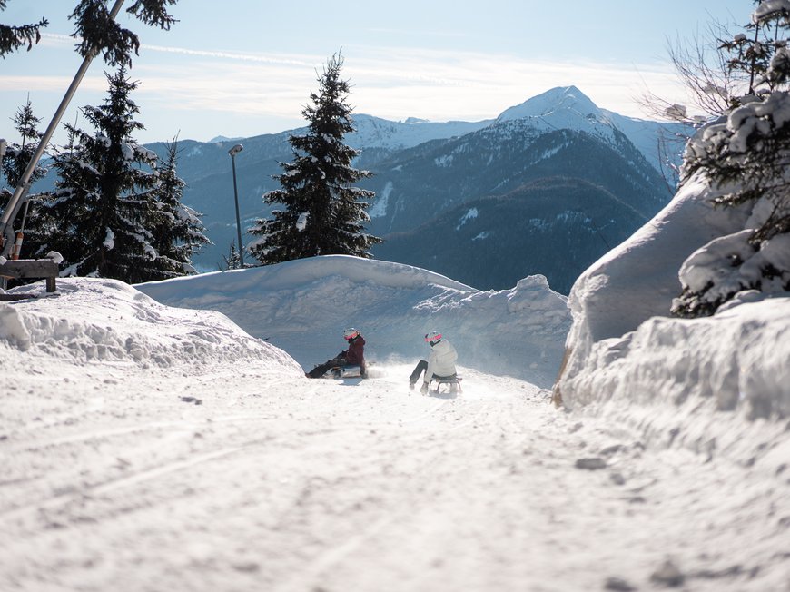 Lahnerhof: Natur- und Wellnesshotel bei Sterzing Zwei Personen rodeln auf einem verschneiten Bergweg mit Tannen und Bergen im Hintergrund