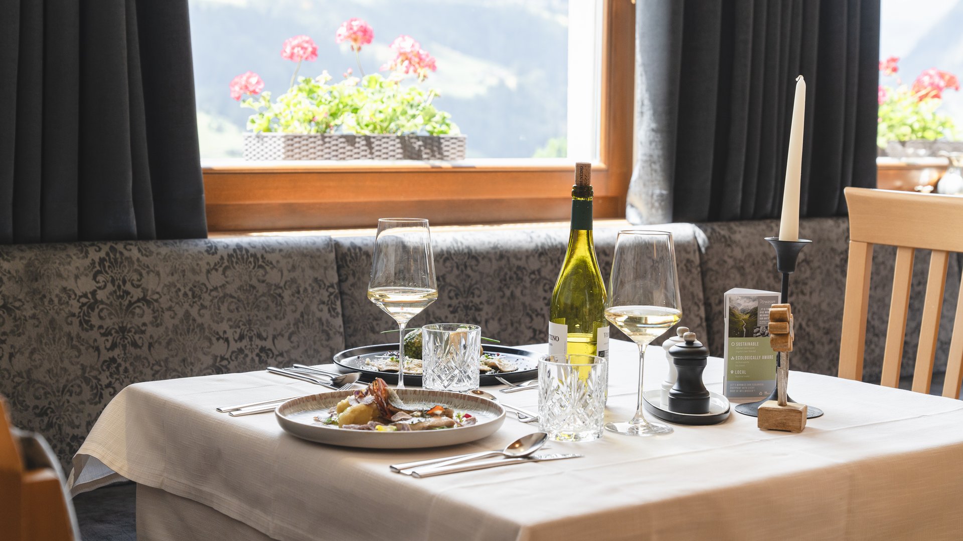 Benvenuti nel ristorante del nostro hotel con spa a Vipiteno Tavolo apparecchiato con vino, candela e piatti vicino a finestra con fiori in ristorante