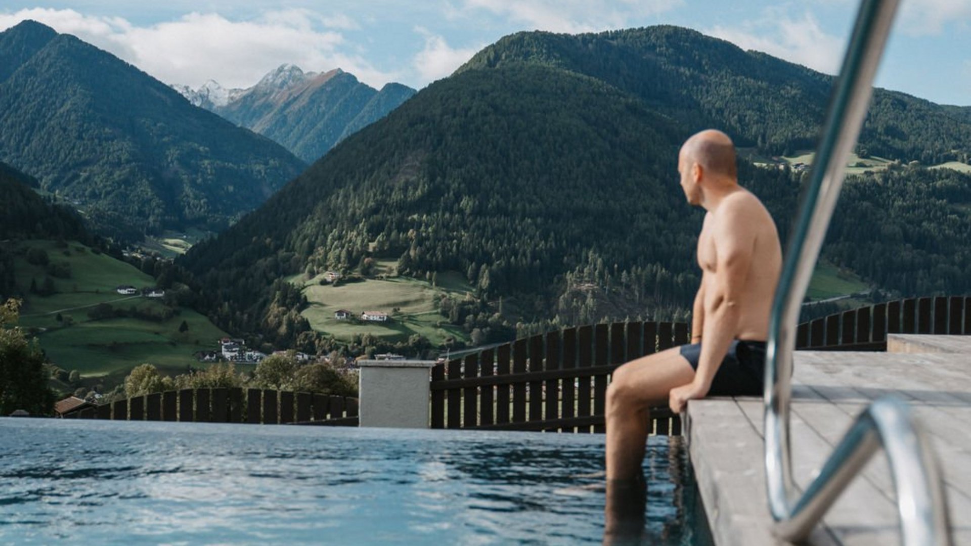 Cercate un hotel a Vipiteno con spa? Benvenuti al Lahnerhof! Uomo seduto a bordo piscina con vista sulle montagne verdi e cielo azzurro