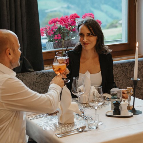 Hotel bei Sterzing für Wellness- und Aktivurlaub Paar stößt mit Getränken in einem Restaurant mit Fensterblick auf Blumen an