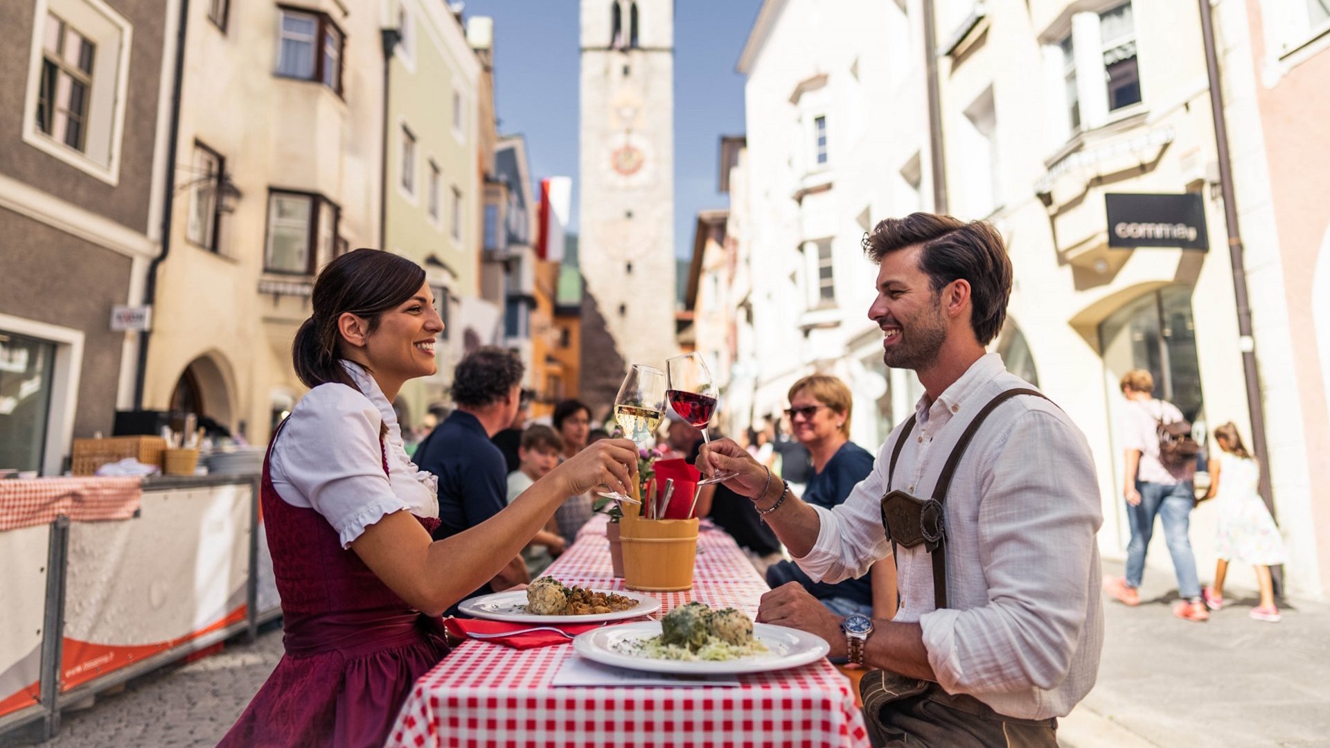 Wanderung auf den Rosskopf und mehr Paar in traditioneller Kleidung stößt mit Weingläsern in einer belebten Straße an