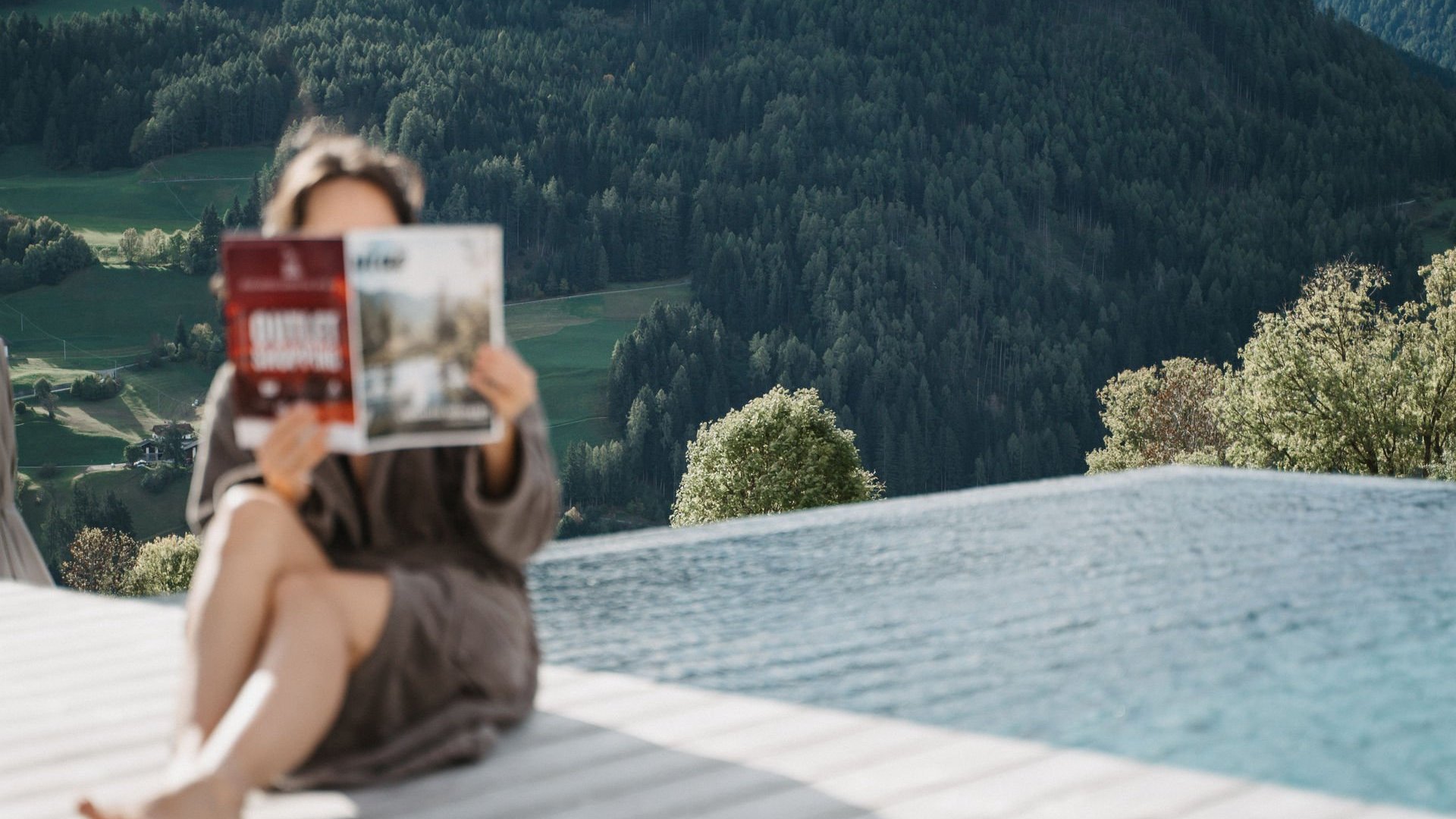 Cercate un hotel a Vipiteno con spa? Benvenuti al Lahnerhof! Persona che legge una rivista vicino alla piscina con foresta e montagne sullo sfondo