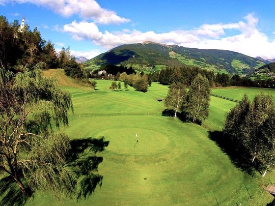 Hiking in Ratschings and surroundings Golf course with green mountains and church under clear blue sky