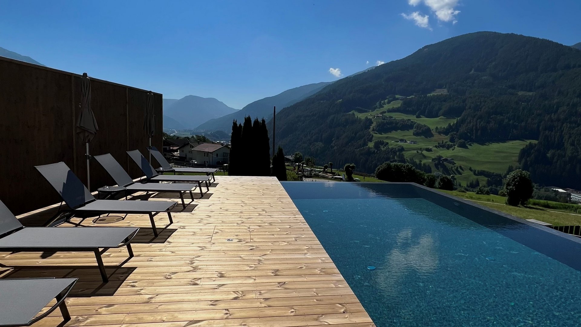 Our hiking and wellness hotel near Sterzing: Lahnerhof Infinity pool with mountain view and lounge chairs on wooden deck under sunny sky