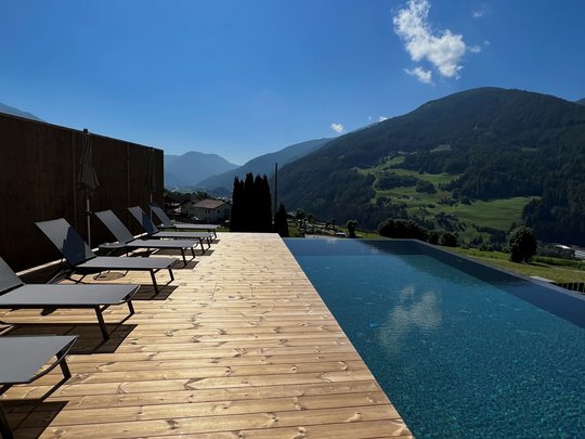 Our hiking and wellness hotel near Sterzing: Lahnerhof Infinity pool with mountain view and lounge chairs on wooden deck under sunny sky