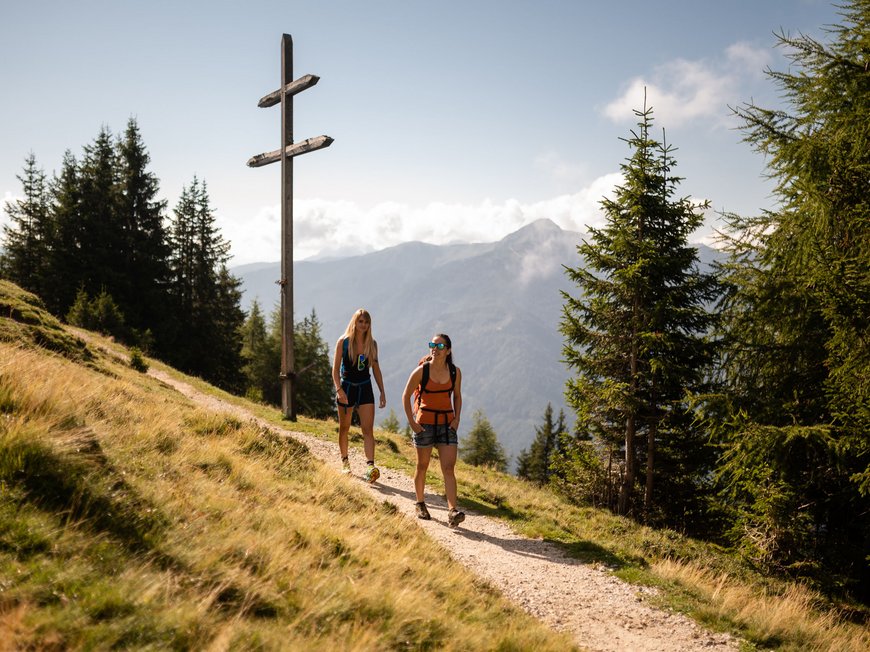 Lahnerhof, Ihr Hotel bei Sterzing Zwei Wanderinnen auf Bergweg neben großem Holzkreuz bei sonnigem Wetter