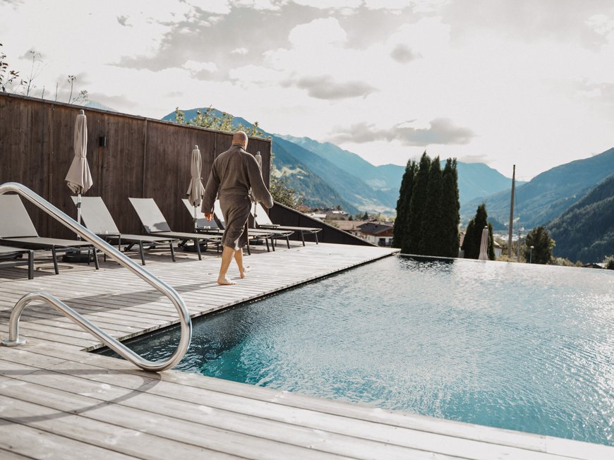 Lahnerhof, your hotel near Sterzing Person in bathrobe walking by pool with mountain view and lounge chairs